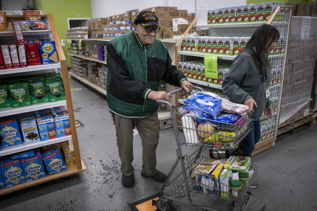 David looks at the camera with a full cart of groceries, volunteer helping guide his cart