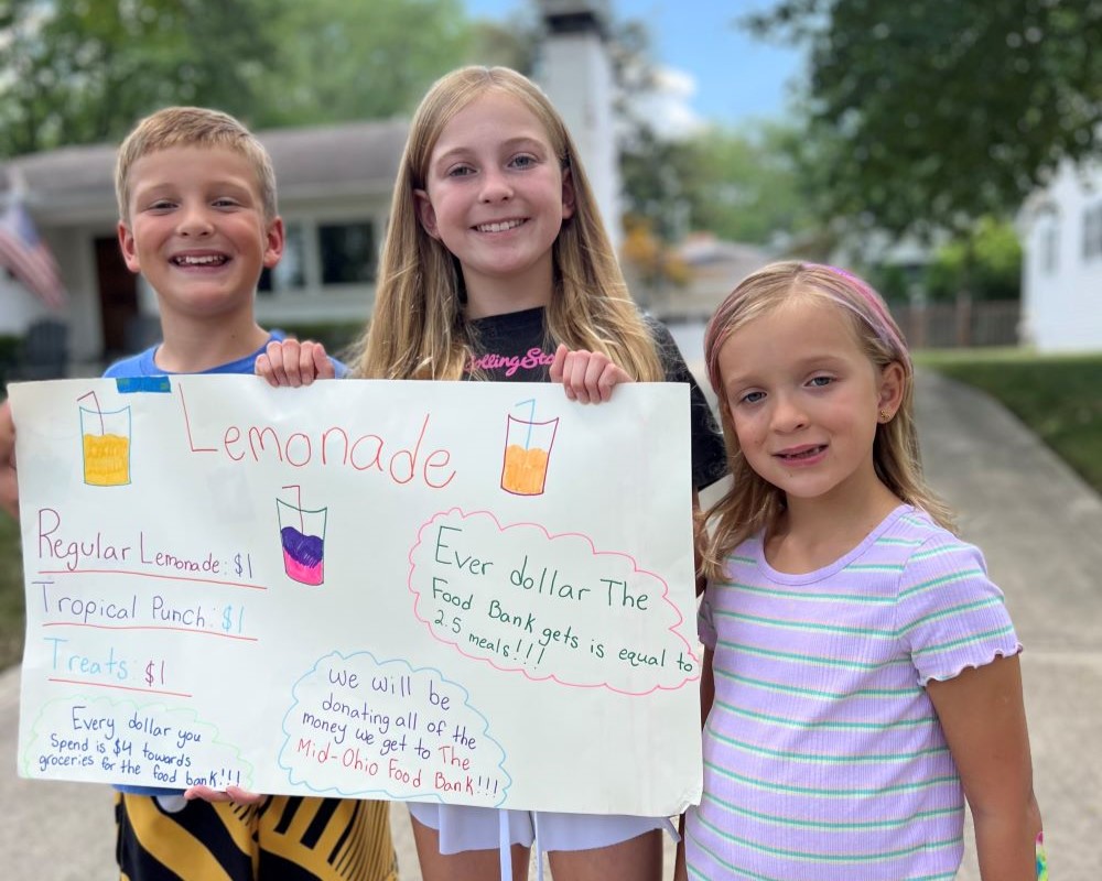 Three children smile holding their lemonade stand sign to raise money for the food bank
