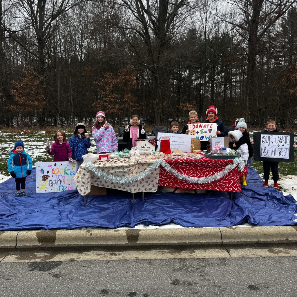 Vinny and his neighborhood friends stand behind their cookie table holding posters and smiling